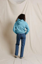 A person standing with their back to the camera, wearing a light blue hooded sweatshirt and blue jeans, posed against a draped off-white fabric backdrop.