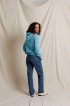 A woman stands with her back to the camera wearing a light blue hoodie and blue jeans, posing in front of a draped off-white fabric backdrop.