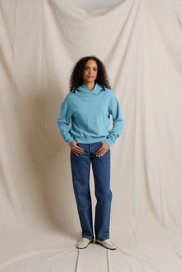 A woman stands against a draped cream fabric backdrop wearing a light blue pullover hoodie, dark blue jeans, and white slip-on shoes.
