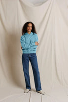 A woman stands against a draped cream fabric backdrop wearing a light blue hoodie, dark blue jeans, and white slip-on shoes.