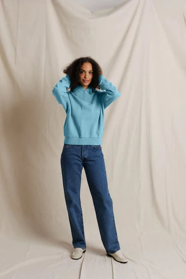 A woman stands against a draped cream fabric backdrop, wearing a light blue sweatshirt, dark blue jeans, and white shoes.