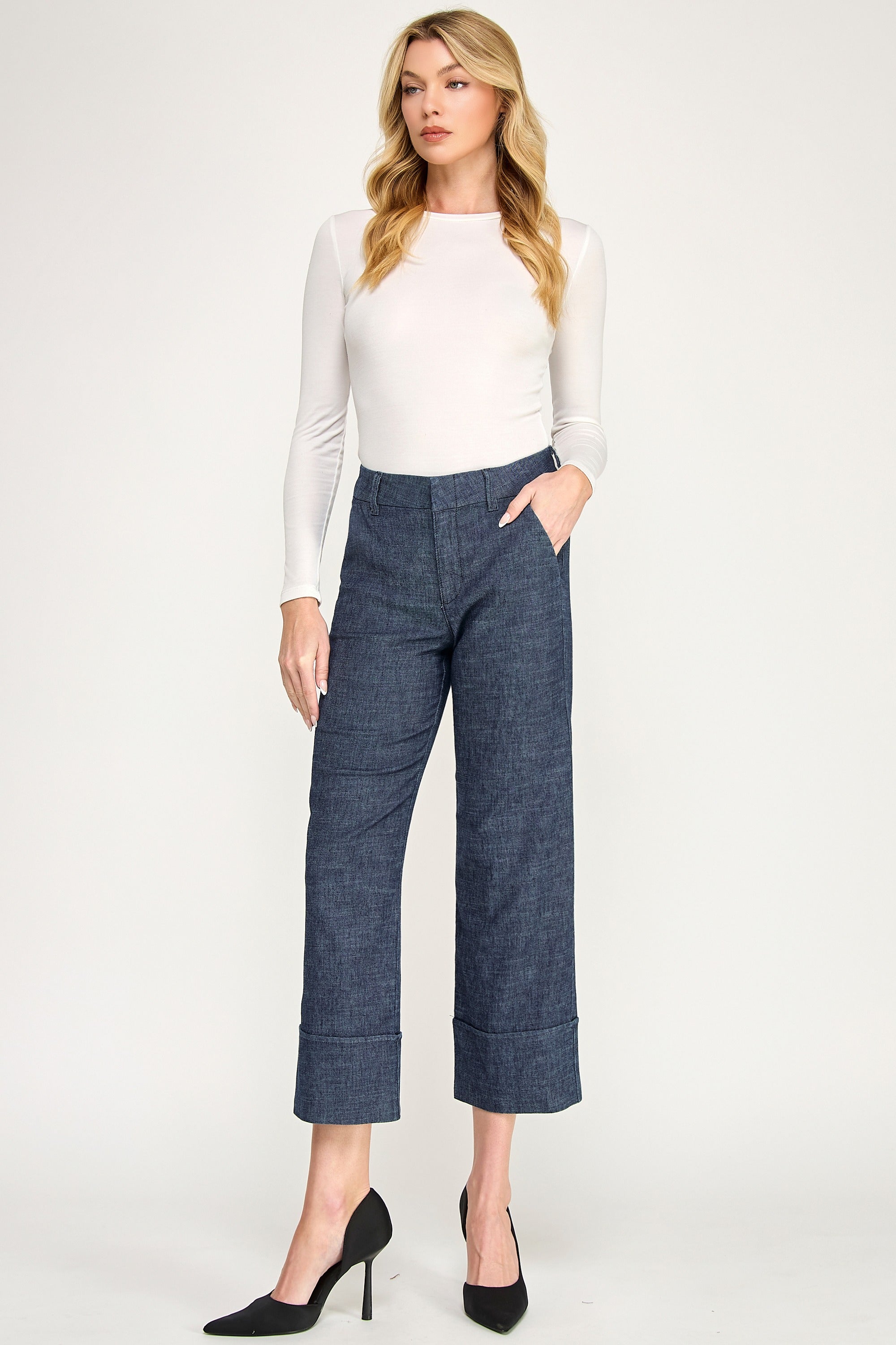 A woman modeling a white long-sleeve mock-neck top and dark blue cropped wide-leg trousers with rolled cuffs, paired with black pointed-toe heels, against a light gray studio background.