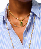 A close-up of a person wearing layered gold necklaces, including a chain with a small green and gold pendant, over a blue and white striped button-down shirt.