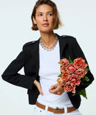 A woman modeling a black cropped military-style jacket over a white t-shirt, accessorized with a chunky silver necklace and a brown belt, while holding a bouquet of pink and red peony flowers against a light gray background.