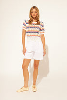 Woman wearing a colorful zigzag pattern top and white shorts on a plain background