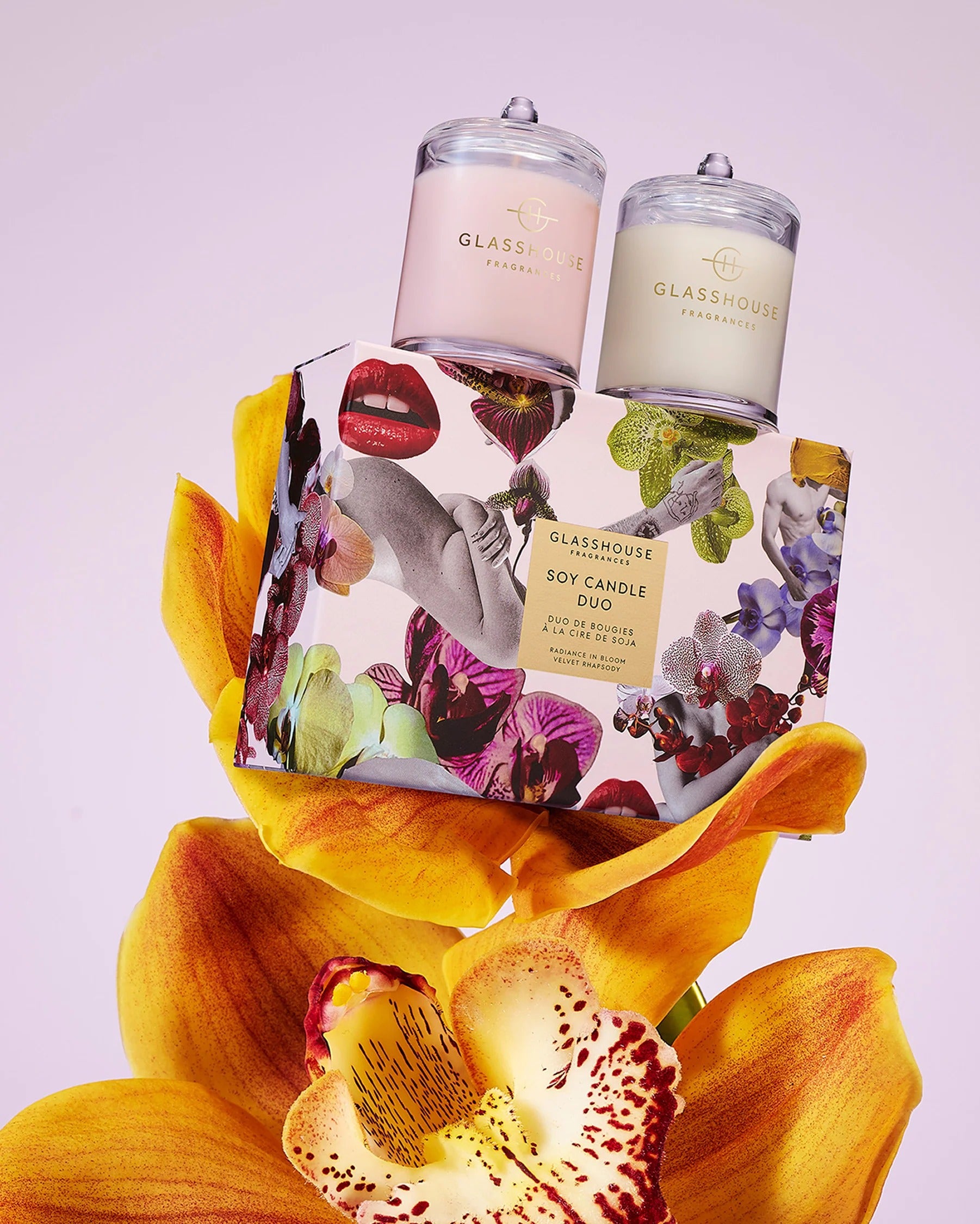 Two glass candles with floral packaging and a large yellow orchid on a light purple background.