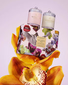 Two glass candles with floral packaging and a large yellow orchid on a light purple background.