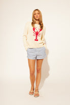 Woman wearing a cream sweater with a red lobster design and blue shorts on a white background