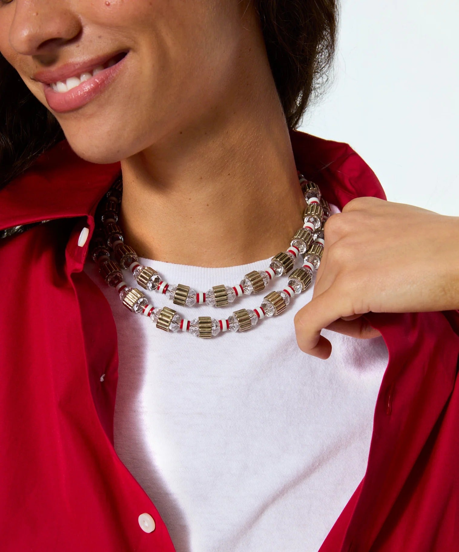 A close-up of a model wearing a double-strand statement necklace made of metallic cylindrical beads with red and white stripes, styled over a white t-shirt and red button-up shirt.