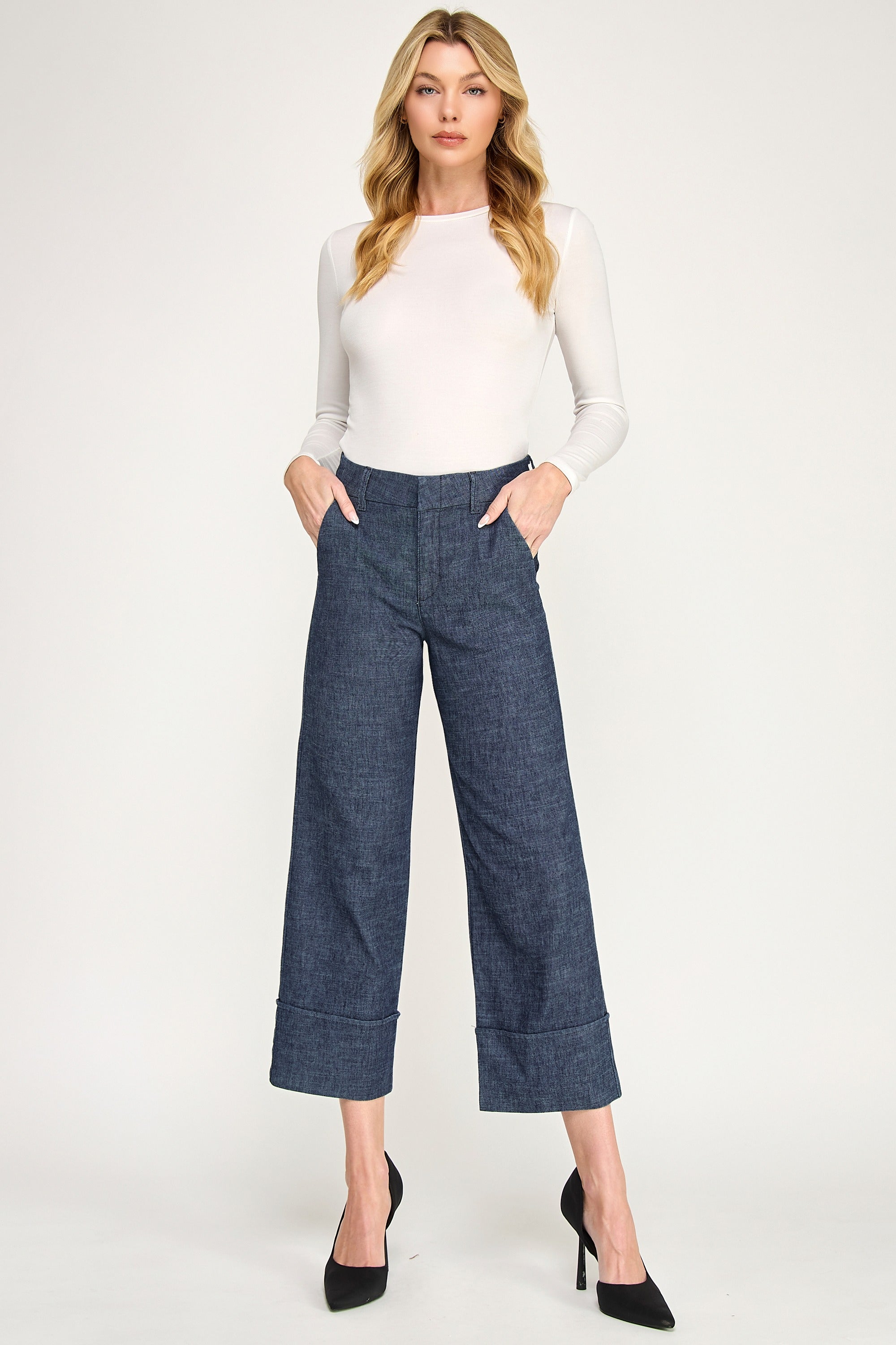 A woman modeling a pair of dark blue cropped wide-leg pants with a subtle textured pattern, paired with a white long-sleeve top and black heels, against a plain light gray studio background.
