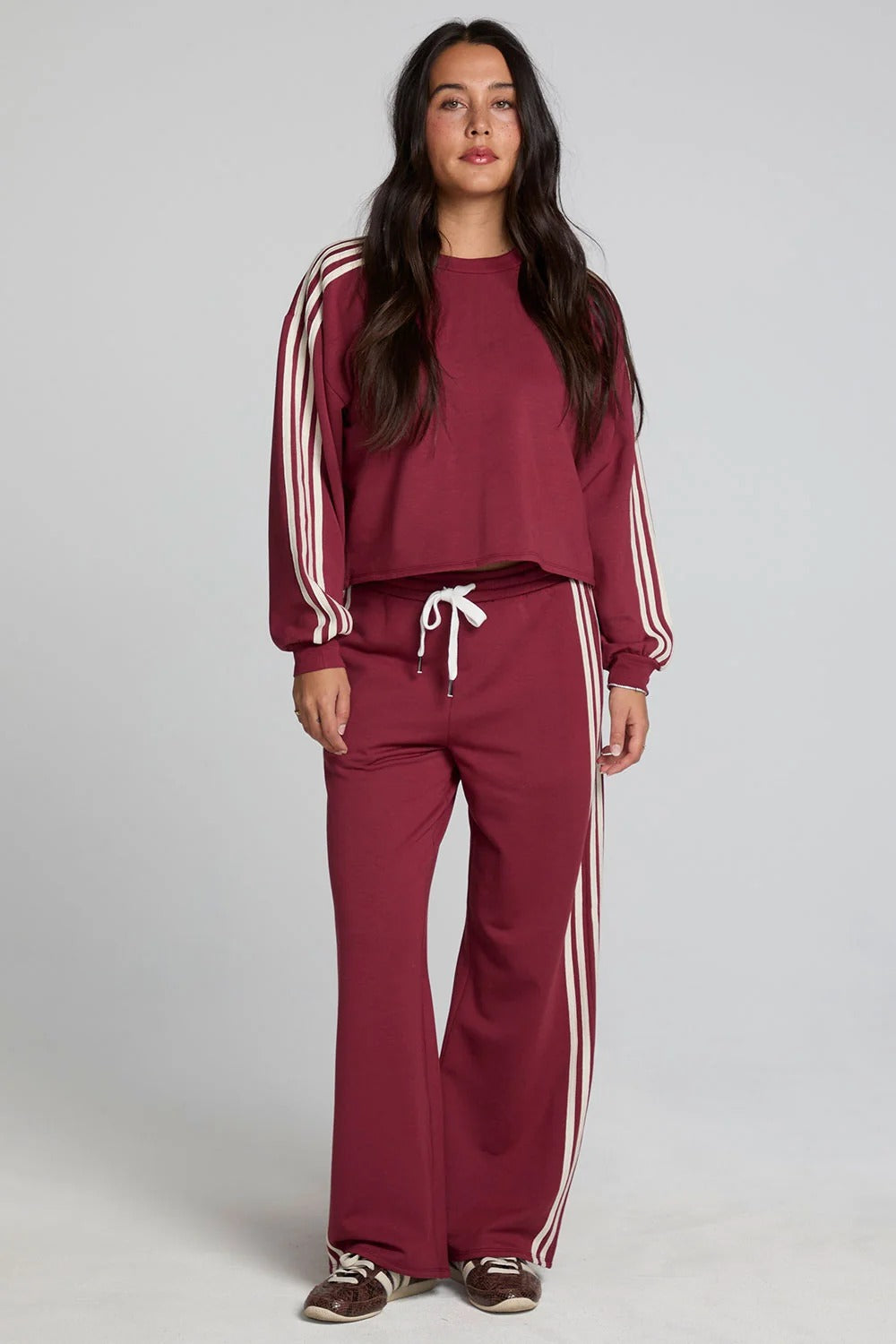 A woman modeling a matching burgundy tracksuit with white stripes on the sleeves and pants, featuring a crewneck sweatshirt and wide-leg sweatpants with a white drawstring, set against a plain light gray studio background.