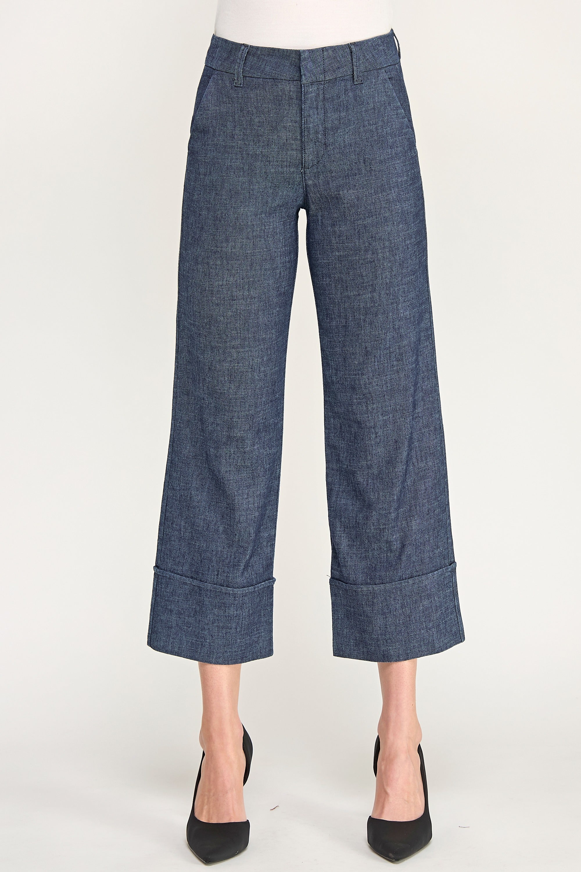 A model is shown from the waist down wearing a pair of dark blue cropped trousers with rolled cuffs and black pointed-toe heels, set against a plain light background.