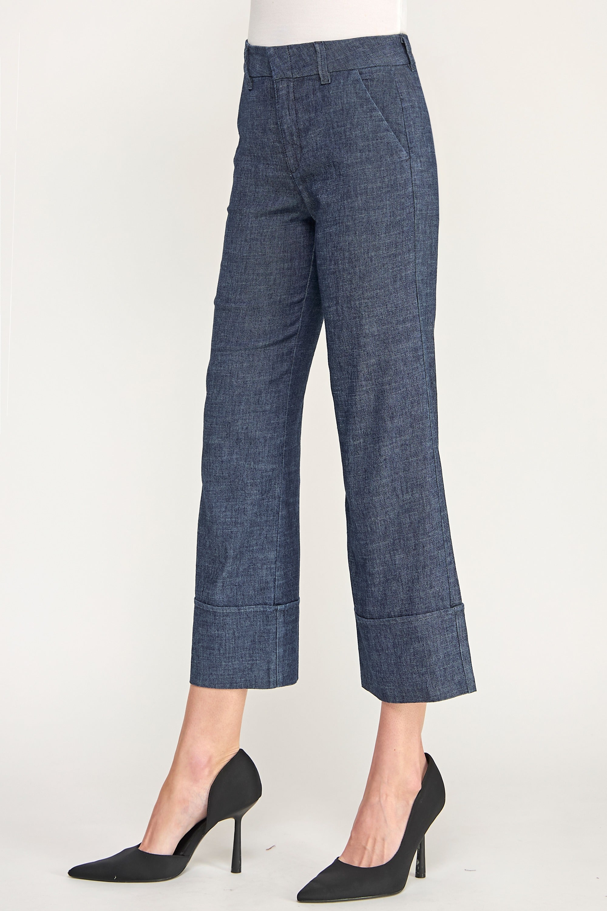 A model is shown from the waist down wearing a pair of dark blue cropped trousers with rolled cuffs and black pointed-toe heels, set against a plain light background.