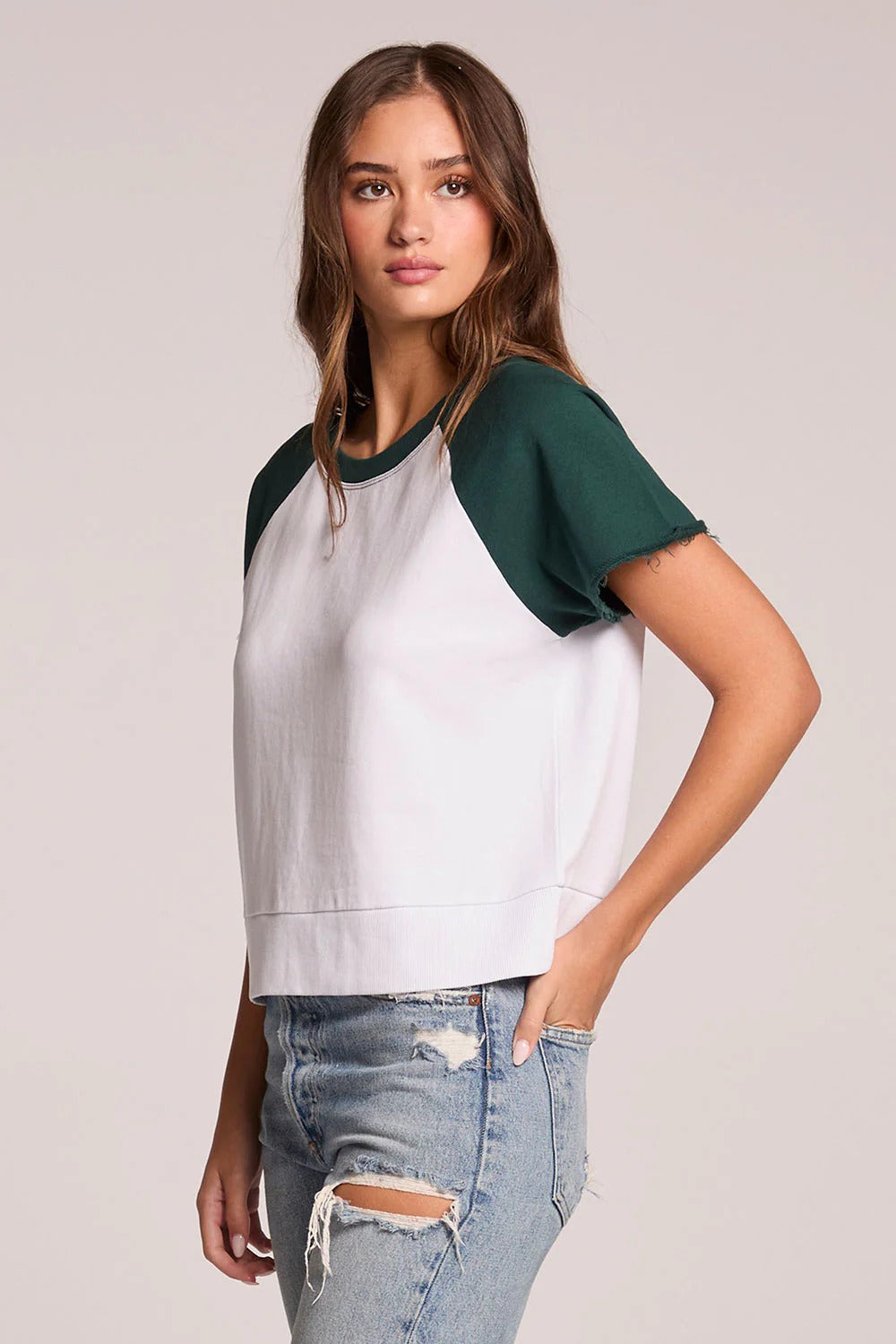 Woman wearing a white and green baseball tee with ripped jeans on a plain background