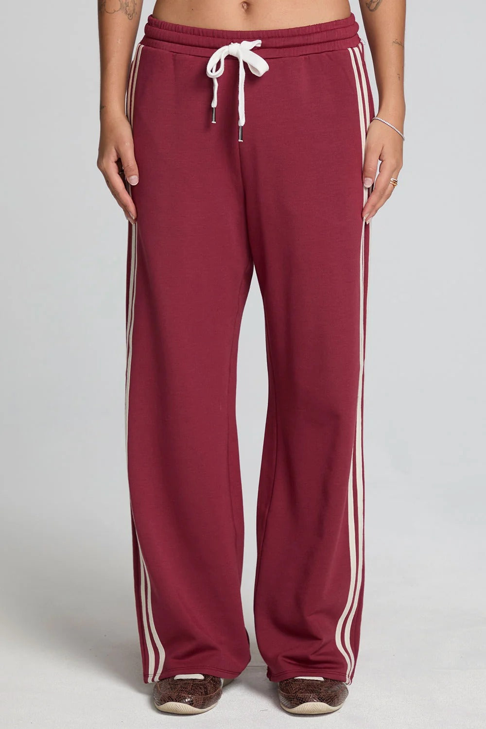 A person wearing burgundy wide-leg sweatpants with white side stripes and a white drawstring, shown from the waist down against a light gray studio background.