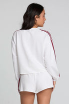 A woman shown from the back wearing a white cropped sweatshirt and matching white shorts, both with red stripes down the sleeves and sides, against a light gray studio background.