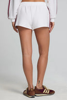 White athletic shorts with burgundy side stripes, worn by a model from the back, paired with brown and gold sneakers against a light gray studio background.