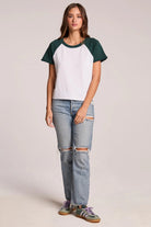 Woman wearing a white and green baseball shirt with ripped jeans on a plain background