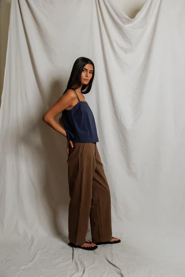 Woman wearing a navy top and brown pants against a white draped curtain background
