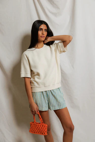 Woman wearing a beige sweater and striped shorts, holding an orange bag against a plain background