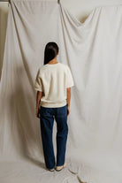 Person wearing a white shirt and blue jeans standing in front of a plain white curtain.