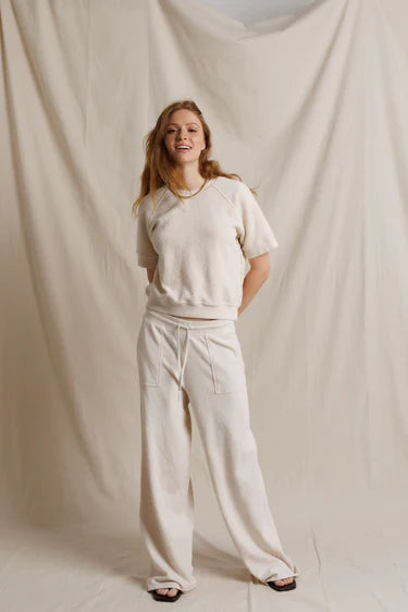 A young woman stands against a draped cream fabric backdrop, wearing a matching cream lounge set consisting of a short-sleeve cropped sweatshirt and wide-leg sweatpants.