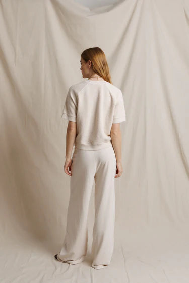 A woman stands with her back to the camera wearing a cream-colored short-sleeve top and matching wide-leg trousers, set against a draped cream fabric backdrop.