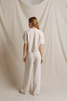 A woman stands with her back to the camera wearing a cream-colored short-sleeve top and matching wide-leg trousers, set against a draped cream fabric backdrop.