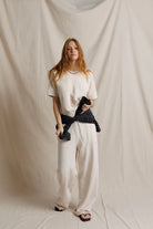 A woman stands against a draped cream fabric backdrop wearing a cream short-sleeve knit top, matching cream wide-leg pants, and black sandals. She has a dark navy plaid shirt tied around her waist.