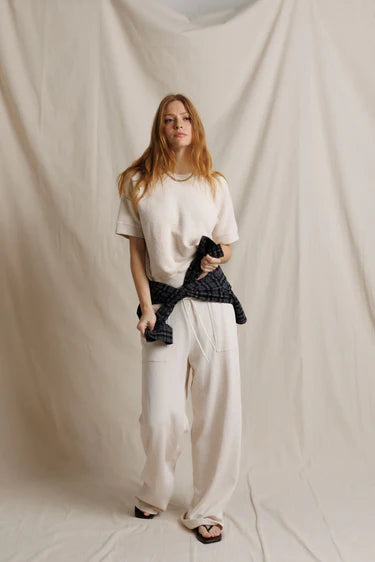 A woman stands against a draped cream fabric backdrop wearing a cream short-sleeve knit top, matching cream wide-leg pants, and black sandals. She has a dark navy plaid shirt tied around her waist.