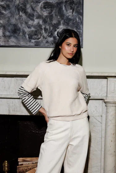 Woman wearing a white sweater with striped sleeves and white pants standing in front of a marble fireplace.