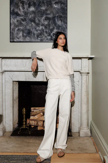 A woman models a cream-colored knit crewneck sweater and matching wide-leg trousers, styled with a striped long-sleeve shirt underneath and brown leather sandals, standing in front of a white marble fireplace.