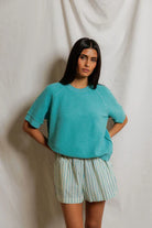 Woman wearing a teal sweater and striped shorts against a plain background