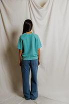 Person wearing a teal shirt and blue jeans standing against a plain white background