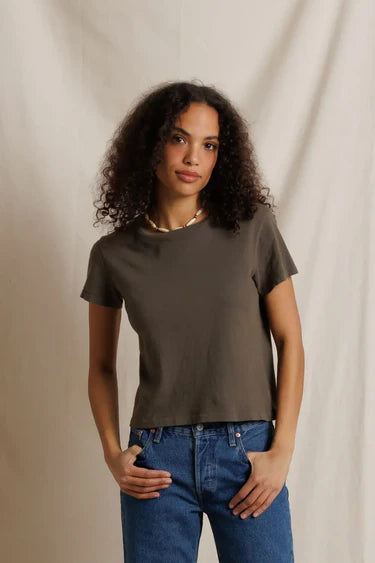 A woman with curly dark hair wearing a dark brown short-sleeve crewneck t-shirt and blue jeans, standing against a draped beige fabric backdrop.