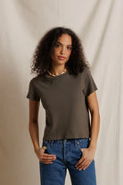 A woman with curly dark hair wearing a dark brown short-sleeve crewneck t-shirt and blue jeans, standing against a draped beige fabric backdrop.
