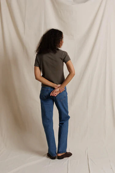 A person standing with their back to the camera, wearing a dark brown short-sleeve t-shirt, blue jeans, and brown shoes, posed against a draped off-white fabric backdrop.