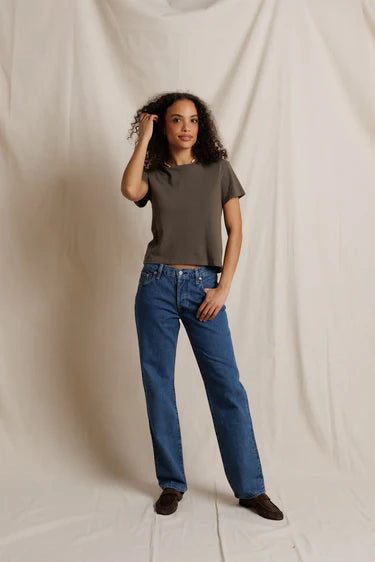 A woman modeling a dark olive green short-sleeve crop top and blue straight-leg jeans, standing against a draped off-white fabric backdrop.