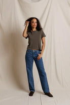 A woman modeling a dark olive green short-sleeve crop top and blue straight-leg jeans, standing against a draped off-white fabric backdrop.