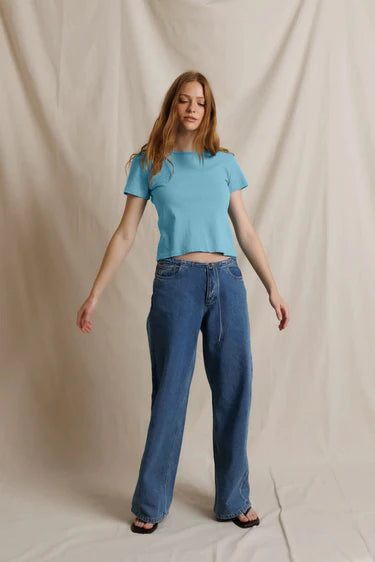 A woman modeling a light blue short-sleeve crop top and wide-leg blue jeans against a draped beige fabric backdrop.