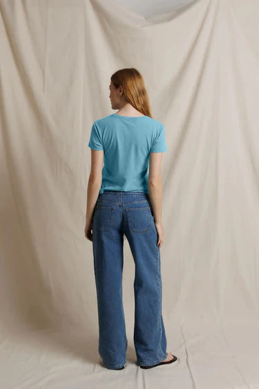A woman stands with her back to the camera wearing a light blue short-sleeve t-shirt and wide-leg blue jeans, standing against a draped beige fabric backdrop.