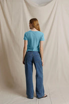 A woman stands with her back to the camera wearing a light blue short-sleeve t-shirt and wide-leg blue jeans, standing against a draped beige fabric backdrop.