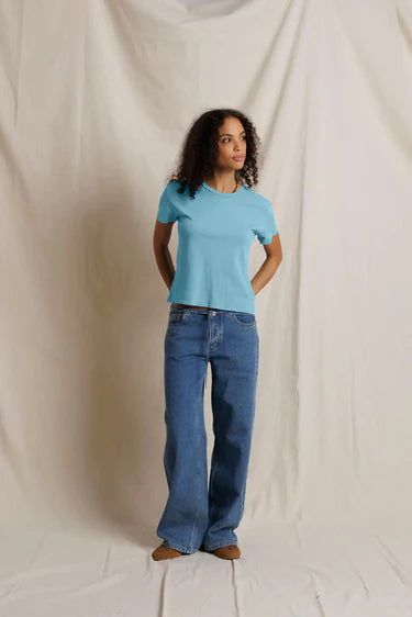 A woman modeling a light blue short-sleeve crewneck t-shirt and wide-leg blue jeans, standing against a draped off-white fabric backdrop.