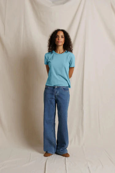 A woman stands against a draped cream fabric backdrop wearing a light blue short-sleeve crewneck t-shirt and wide-leg blue jeans.