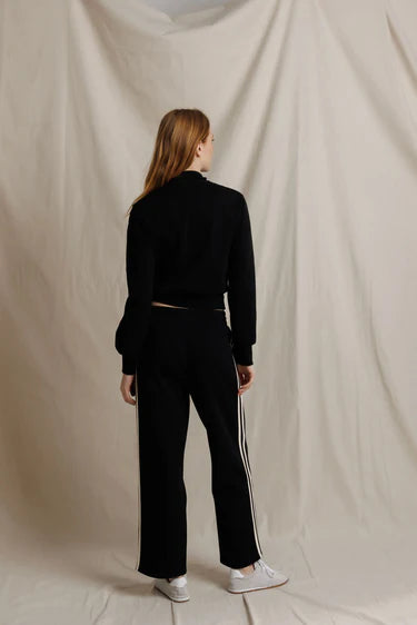 A woman is shown from behind wearing a black knit long-sleeve crop top and matching black wide-leg trousers with white side stripes, paired with white sneakers, set against a draped beige fabric backdrop.