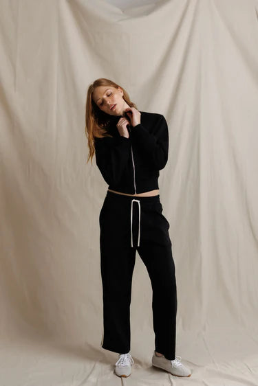 A woman modeling a black cropped zip-up jacket and matching black sweatpants with a white drawstring, paired with white sneakers, posed against a draped beige fabric backdrop.