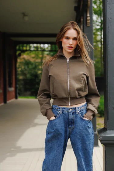Woman wearing a brown jacket and blue jeans standing outdoors.