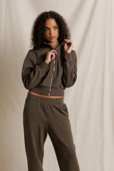 A woman modeling a matching dark brown cropped zip-up hoodie and sweatpants set against a draped beige fabric backdrop.