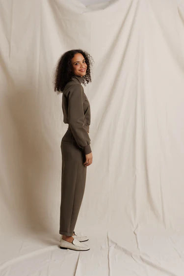 A woman with curly dark hair stands in profile wearing a matching taupe-brown long-sleeve top and slim trousers, paired with white loafers, against a draped off-white fabric backdrop.