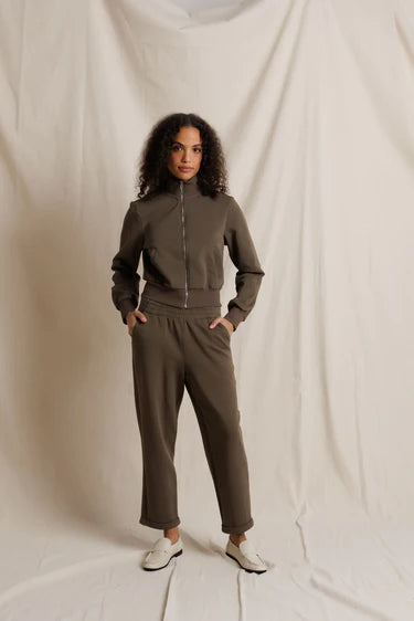 A woman modeling a matching two-piece tracksuit in a muted olive-brown color, consisting of a zip-up long-sleeve top and matching pants, standing against a draped off-white fabric backdrop.